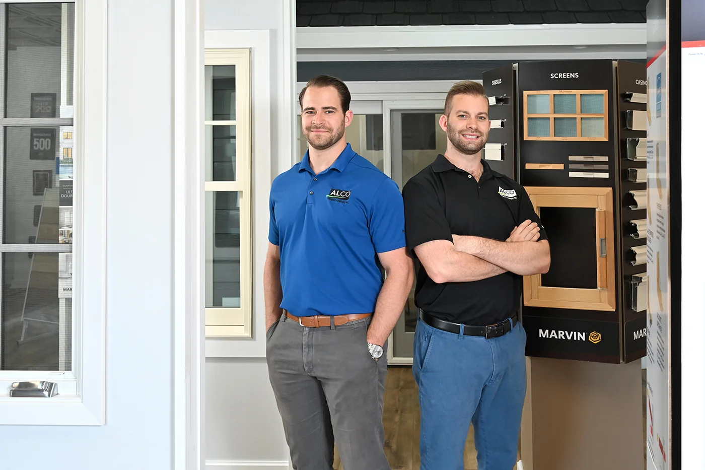 Alco Products&rsquo; owners, two brothers in Alco polo shirts, standing in the North Bethesda showroom beside a Marvin window display