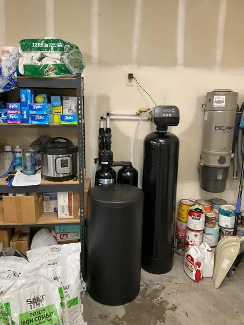 A Kinetico softener and brine tank installed in the corner of a garage beside shelving and salt bags